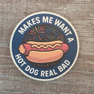 以下が含まれることがあります： 青い縁と白い文字「MAKES ME WANT A HOT DOG REAL BAD」が入った丸いコースター。中央には、花火とホットドッグのグラフィックが描かれています。
