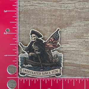 May include: A sticker featuring a skeleton in a boat, holding an oar and an American flag. The sticker has the text "Undefeated Since 1776" on a banner. The sticker is on a wooden surface.