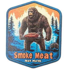 May include: A Bigfoot-themed sticker with a hexagonal shape. The illustration features a hairy Bigfoot figure grilling meat over a barbecue. The background shows a forest scene with orange and blue hues. The text "Smoke Meat Not Meth" is displayed.