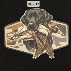 Puede incluir: Una pegatina que muestra a un labrador retriever negro sosteniendo un pato real en su boca. El perro y el pato están centrados sobre un fondo hexagonal con un patrón de camuflaje en tonos beige, marrón y gris. El texto DU-012 aparece arriba.