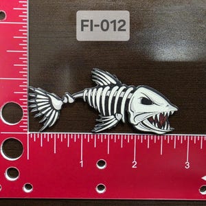 May include: A white and black fish skeleton patch with red teeth. The patch has a white background and the text "FI-012" is printed on a white rectangle.