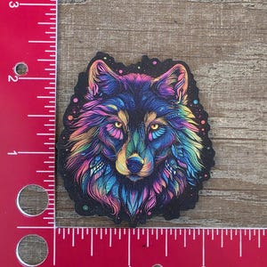 May include: A colourful sticker of a wolf's head. The wolf has a black background with vibrant rainbow-coloured fur, including shades of blue, purple, yellow, and green. The sticker is approximately 6.35 cm wide.