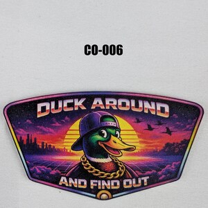 Puede incluir: Un parche colorido con un pato de dibujos animados que lleva una gorra morada y una cadena de oro. El parche dice "DUCK AROUND AND FIND OUT" con un fondo de puesta de sol y horizonte de la ciudad. El parche está etiquetado como "CO-006".