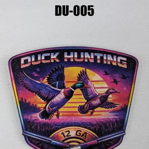 May include: A colorful graphic featuring two ducks in flight over a sunset landscape. The design includes the text "DUCK HUNTING" and "12 GA" with crossed shotguns. The image has a retro, vintage style.