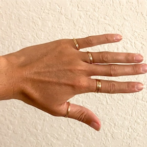 May include: A hand with five fingers, each adorned with a thin, gold ring. The rings are simple bands, some with a double band design. The skin tone is light, and the background is a textured white wall. The rings are likely jewelry.