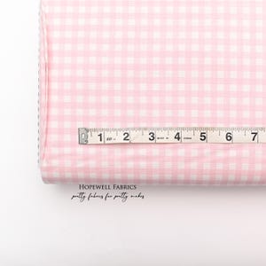May include: Pink and white gingham fabric with a measuring tape. The fabric is folded over, showing the pattern and texture. The measuring tape shows inches. The text "HOPEWELL FABRICS" and "pretty fabrics for pretty makes" is visible.