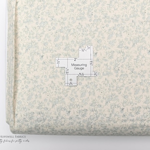 May include: A roll of fabric with a delicate floral pattern in shades of cream and light blue. A measuring gauge is placed on top, with measurements in inches. The fabric is labeled "HOPEWELL FABRICS."