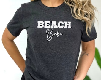 May include: A dark grey heather t-shirt with the text "Beach Babe" in white lettering. The text is in a bold, blocky font with the word "Babe" in a cursive script.