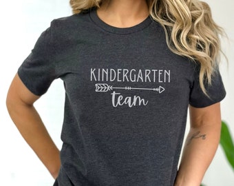 May include: A dark heather grey t-shirt with a white arrow and the text "Kindergarten team" printed on the front.