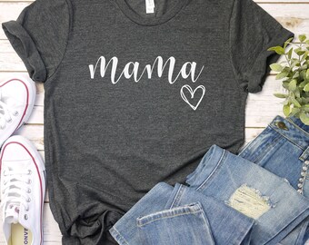 May include: A dark gray t-shirt with the word "mama" written in white cursive lettering and a white heart drawn next to it. The shirt is laid flat on a wooden surface with a pair of white sneakers and a pair of blue jeans.
