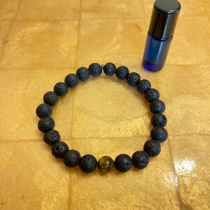 以下が含まれることがあります： 黒い溶岩のビーズで作られたブレスレットに、タイガーアイのビーズが1つ付いている。ブレスレットの隣には、黒いキャップが付いた小さな青いガラス瓶がある。