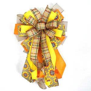 May include: A large, multi-layered bow made with yellow, orange, and brown ribbon. The ribbon has a plaid pattern and a sunflower print. The bow is perfect for a fall or autumn wreath.