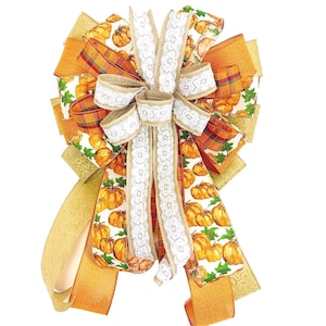 May include: A large, decorative bow made with multiple ribbons. The bow features a white ribbon with lace trim, a brown burlap ribbon, and a ribbon with a pumpkin print. The bow is tied with a gold ribbon.