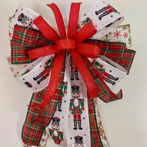 May include: A festive Christmas bow featuring red, white, and green ribbons. The bow has a central red ribbon and is adorned with ribbons printed with nutcrackers, plaid patterns, and snowflakes. Perfect for holiday decorating.