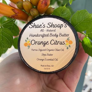 May include: A round container of Shae's Shoap's Handcrafted Body Butter, Orange Citrus. The label lists ingredients: Yarrow Infused Organic Olive Oil, Shea Butter, and Orange Essential Oil. The container is held near orange flowers and green leaves.