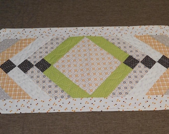 May include: A white table runner with a geometric pattern in shades of green, gray, black, and yellow. The runner has a diamond shape in the center and a scalloped edge.