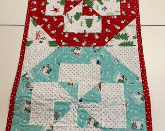 May include: A red, white, and blue Christmas table runner with a snowflake pattern. The runner is made of fabric and has a quilted design. The runner is decorated with a variety of Christmas-themed images, including snowmen, trees, and snow globes.