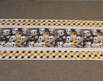 May include: A white table runner with a black and yellow checkered border. The center of the runner features a repeating pattern of white skulls and orange flowers on a black background.