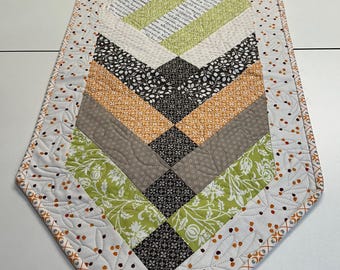 May include: A quilted table runner with a geometric pattern in shades of green, brown, orange, and white. The runner has a scalloped edge with a white background and a small orange and white pattern.