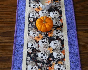 May include: A purple and white table runner with a black background featuring a skull and floral pattern. Three orange pumpkins and a white cup with a candle are placed on the runner.