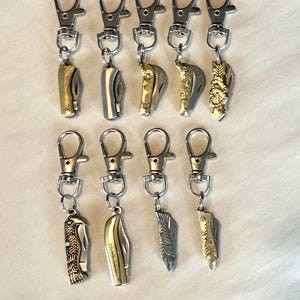 May include: A collection of pocket knife keychains with silver-tone clasps. The knives have various designs, including gold and silver finishes, and decorative patterns. The keychains are arranged against a white background.