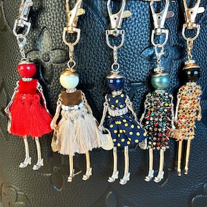 May include: A collection of five doll-shaped keychains. Each keychain features a beaded head, a dress, and metal legs and arms. The dresses are in various colors and styles, including red fringe, navy blue with yellow polka dots, and a jeweled design.
