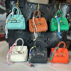 Puede incluir: Una colecci&oacute;n de llaveros de mini bolsos en varios colores, incluyendo turquesa, marr&oacute;n, verde, blanco, negro y naranja. Cada bolso presenta un cierre y asa dorados, con una bufanda decorativa atada. Los llaveros se exhiben frente a un bolso tote negro en relieve.