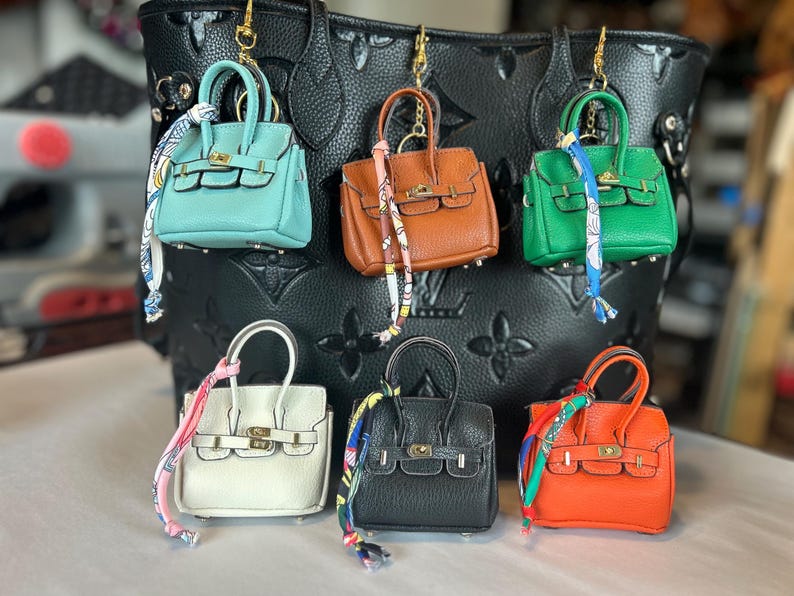 Puede incluir: Una colecci&oacute;n de llaveros de mini bolsos en varios colores, incluyendo turquesa, marr&oacute;n, verde, blanco, negro y naranja. Cada bolso presenta un cierre y asa dorados, con una bufanda decorativa atada. Los llaveros se exhiben frente a un bolso tote negro en relieve.