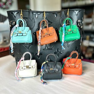 Puede incluir: Una colecci&oacute;n de mini bolsos de mano de cuero en varios colores, incluyendo turquesa, naranja, verde, blanco, negro y marr&oacute;n. Cada bolso tiene un asa superior, un cierre dorado y una bufanda decorativa. Los bolsos cuelgan de un gran bolso tote negro.