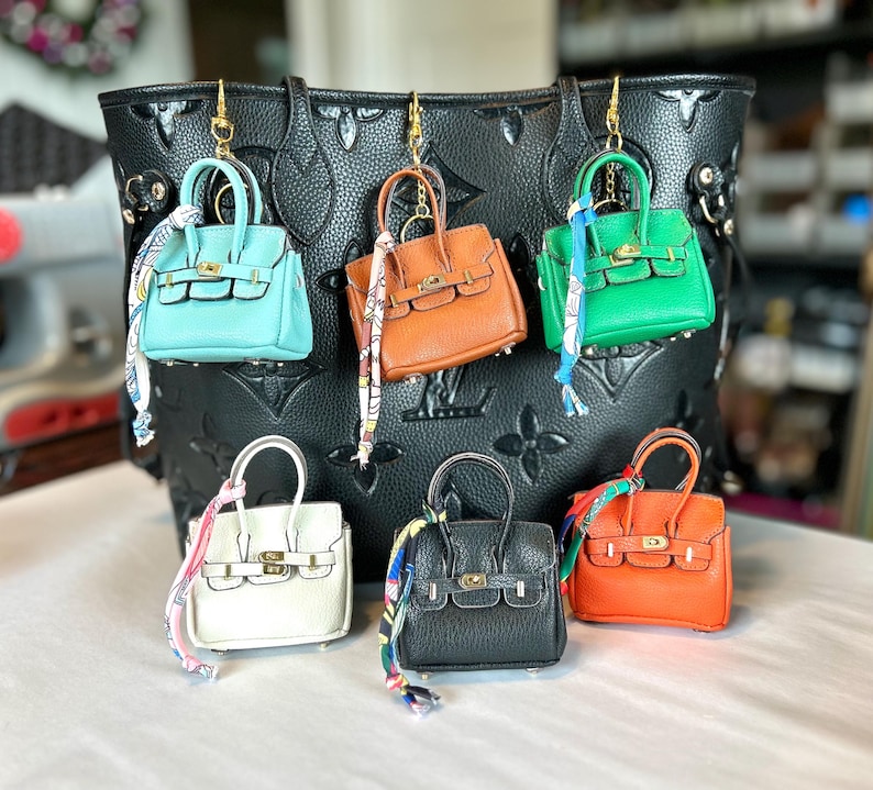 Puede incluir: Una colecci&oacute;n de mini bolsos de mano de cuero en varios colores, incluyendo turquesa, naranja, verde, blanco, negro y marr&oacute;n. Cada bolso tiene un asa superior, un cierre dorado y una bufanda decorativa. Los bolsos cuelgan de un gran bolso tote negro.