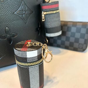 May include: Two black and white plaid zippered pouches with red accents attached to gold keychains. The pouches are hanging from a black leather bag with a monogram pattern.