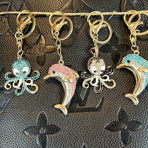 May include: Four keychains with gold-colored hardware. Two keychains feature bejeweled octopus charms in teal and silver. The other two keychains have dolphin charms in pink and teal, also bejeweled. The keychains hang against a black, textured background.
