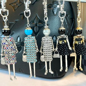 May include: Five silver charm keychains featuring a woman in a different dress. The dresses are turquoise, silver, black, black with white polka dots, and red, blue, and white. Each keychain has a small purse charm.
