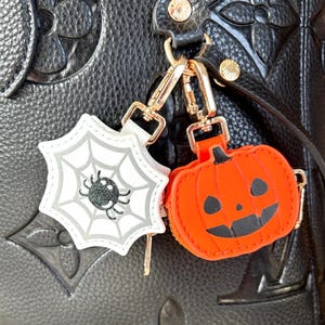 May include: Halloween-themed bag charms. One charm is a white spiderweb with a black spider, and the other is an orange pumpkin with a carved face. Both charms have gold-colored hardware.