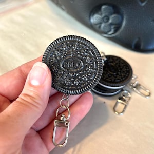 May include: Black Oreo cookie-shaped retractable badge holder with a silver-colored clip. The badge holder has the word "OREO" embossed on the front. Additional Oreo cookies and clips are visible in the background.