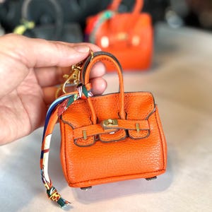 Puede incluir: Llavero de bolso en miniatura naranja con herrajes dorados, un asa superior y un pa&ntilde;uelo decorativo. El peque&ntilde;o bolso tiene una superficie texturizada y un cierre de broche. Otro bolso similar es visible en el fondo.