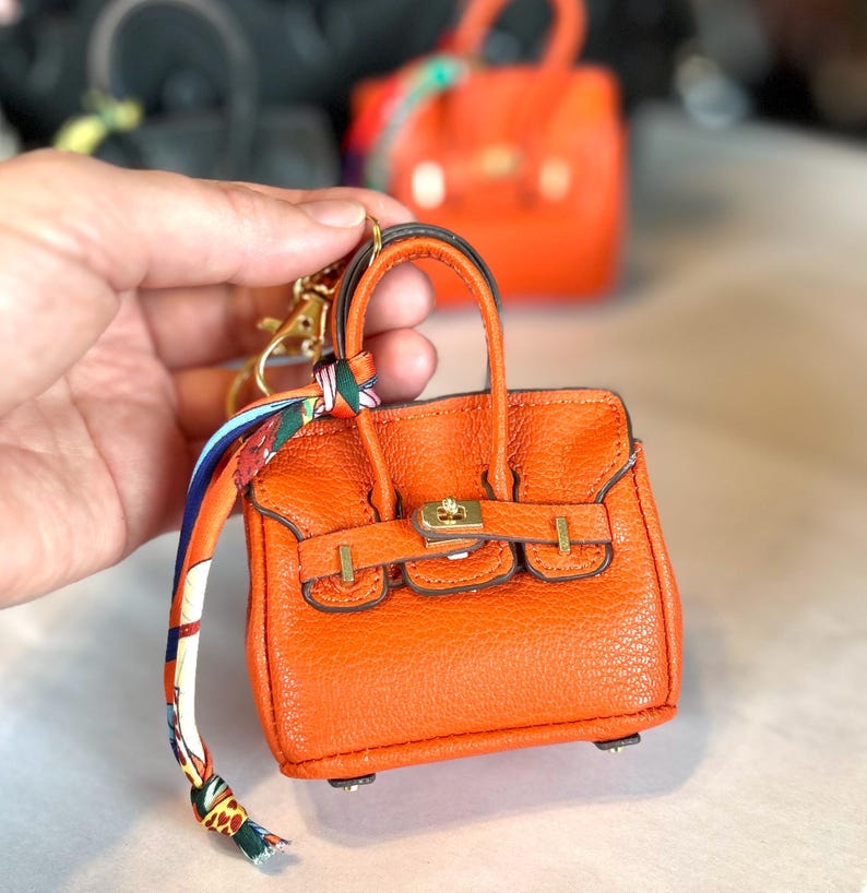 Puede incluir: Llavero de bolso en miniatura naranja con herrajes dorados, un asa superior y un pa&ntilde;uelo decorativo. El peque&ntilde;o bolso tiene una superficie texturizada y un cierre de broche. Otro bolso similar es visible en el fondo.
