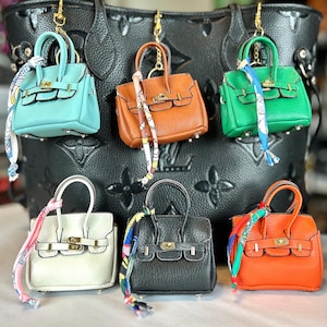 May include: A collection of miniature handbag keychains in various colors, including teal, brown, green, white, black, and orange. Each bag features a gold-tone clasp and a decorative scarf. The keychains are hanging in front of a large black handbag.
