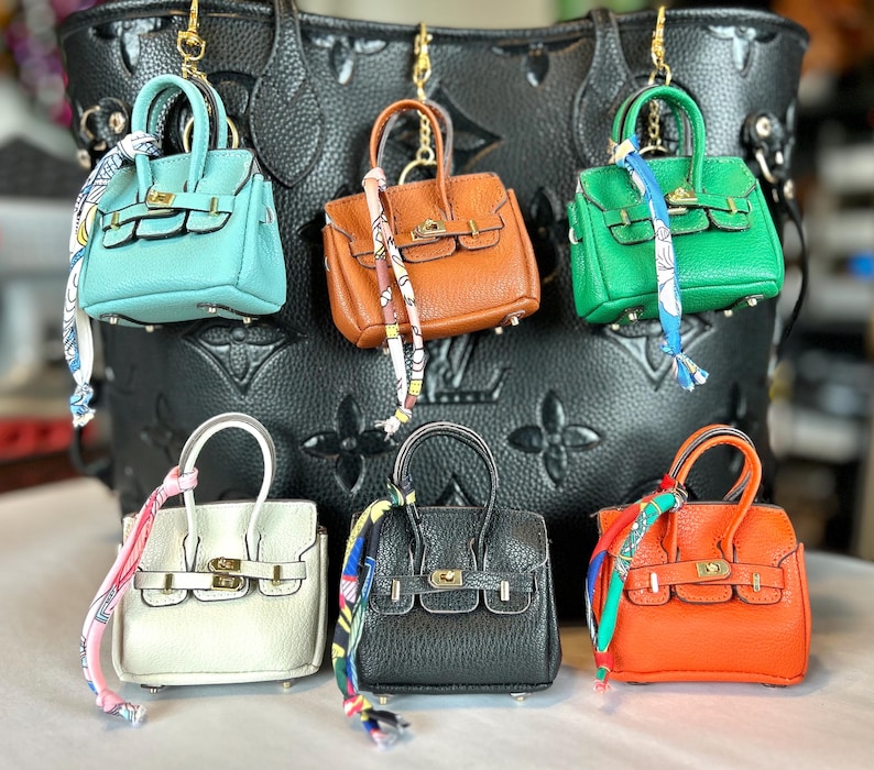 Puede incluir: Una colecci&oacute;n de llaveros de mini bolsos en varios colores, incluyendo turquesa, marr&oacute;n, verde, blanco, negro y naranja. Cada bolso presenta un cierre dorado y una bufanda decorativa. Los llaveros cuelgan frente a un gran bolso negro.