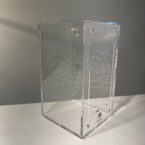 May include: A clear, rectangular acrylic terrarium with a hinged door and ventilation holes. The enclosure features a series of drilled holes in a circular pattern on the side. The bottom right corner has a small logo.