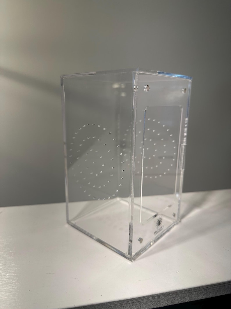 May include: A clear, rectangular acrylic terrarium with a hinged door and ventilation holes. The enclosure features a series of drilled holes in a circular pattern on the side. The bottom right corner has a small logo.