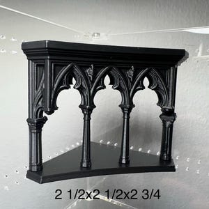 May include: Black corner shelf with gothic arch design. The shelf is 2 1/2 x 2 1/2 x 2 3/4 inches. The shelf has a triangular shape and is made of a solid black material. The shelf has a decorative gothic arch design.