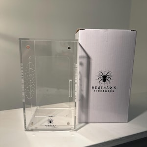 May include: A clear acrylic terrarium with a hinged door and a white cardboard box. The box features a black spider graphic and the text "HEATHER'S HIDEAWAYS". The terrarium is designed for small pets.
