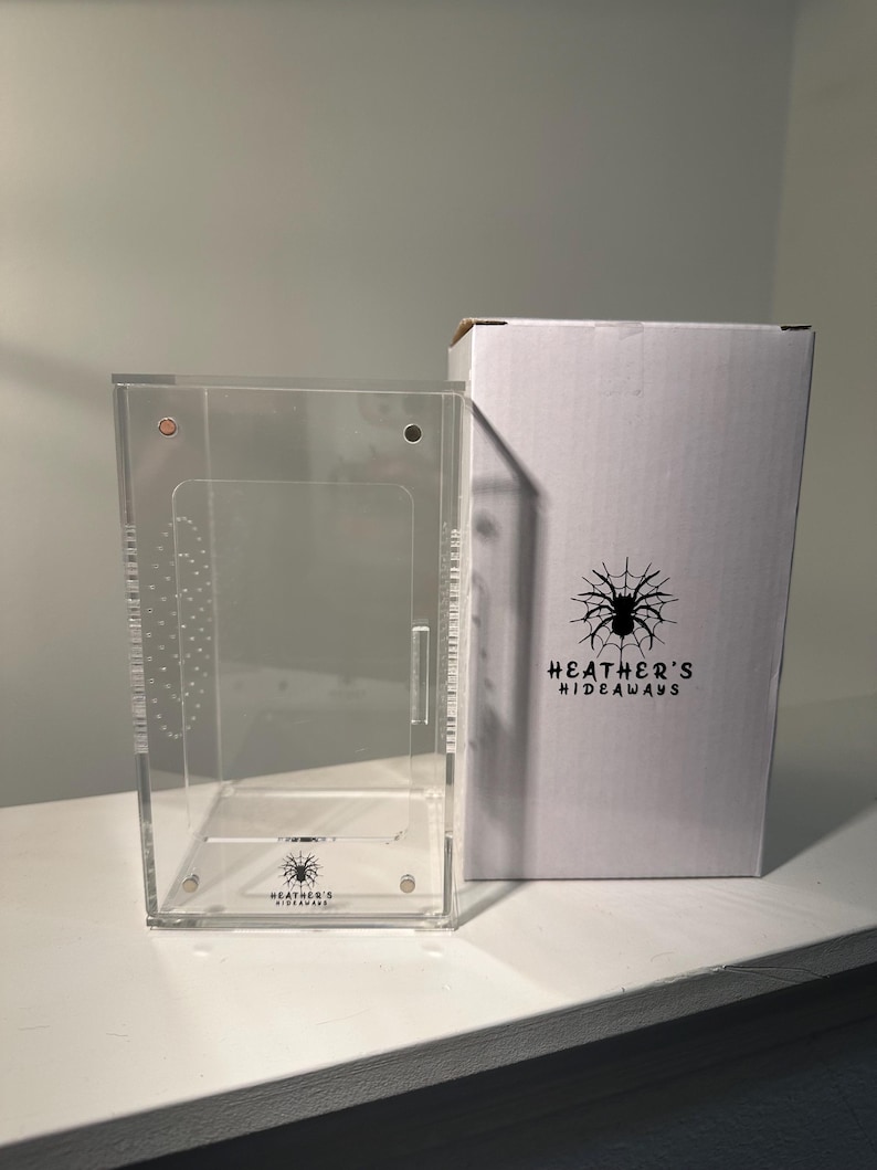 May include: A clear acrylic terrarium with a hinged door and a white cardboard box. The box features a black spider graphic and the text "HEATHER'S HIDEAWAYS". The terrarium is designed for small pets.