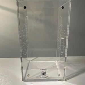 May include: A clear acrylic terrarium with a rectangular shape. The enclosure features small ventilation holes and a hinged lid. The base of the terrarium has the text "HEATHER'S HIDEAWAY" with a spider graphic. The terrarium is designed for small pets.