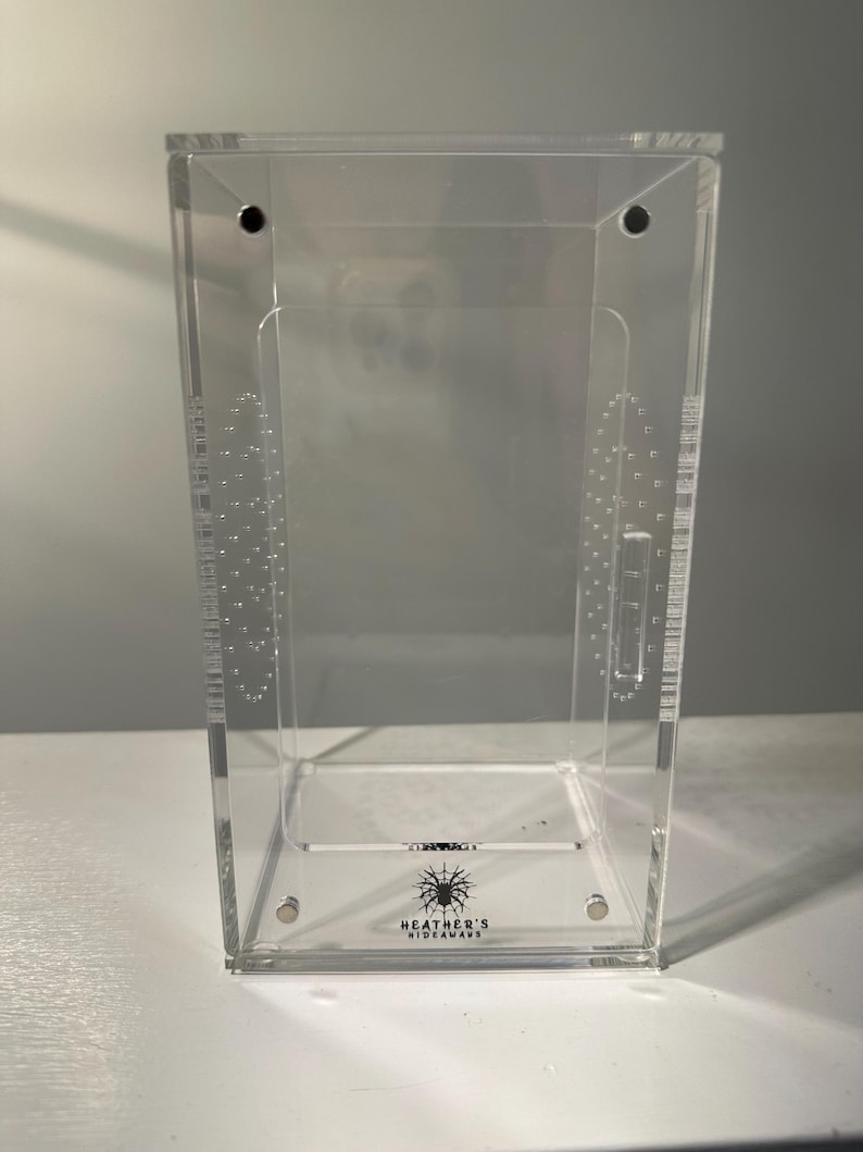 May include: A clear acrylic terrarium with a rectangular shape. The enclosure features small ventilation holes and a hinged lid. The base of the terrarium has the text "HEATHER'S HIDEAWAY" with a spider graphic. The terrarium is designed for small pets.
