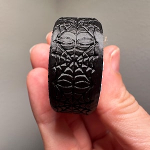 May include: A black ring with a detailed spiderweb design. The ring is made of a solid material and features a repeating pattern of webs and geometric shapes. The ring is held by a hand.