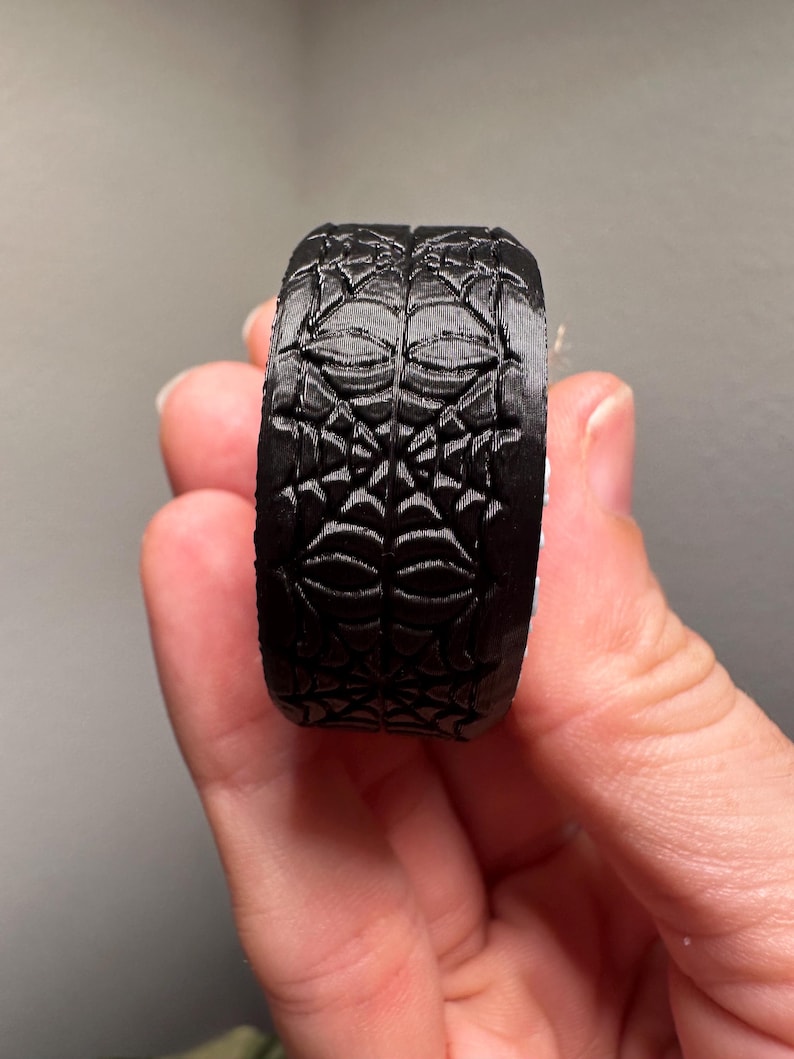 May include: A black ring with a detailed spiderweb design. The ring is made of a solid material and features a repeating pattern of webs and geometric shapes. The ring is held by a hand.