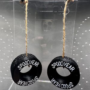 May include: Two black, tire-shaped ornaments with the text "SPOOD YEAR" in white lettering. Each ornament hangs from a natural-colored twine. The ornaments are suspended in front of a clear, reflective surface.