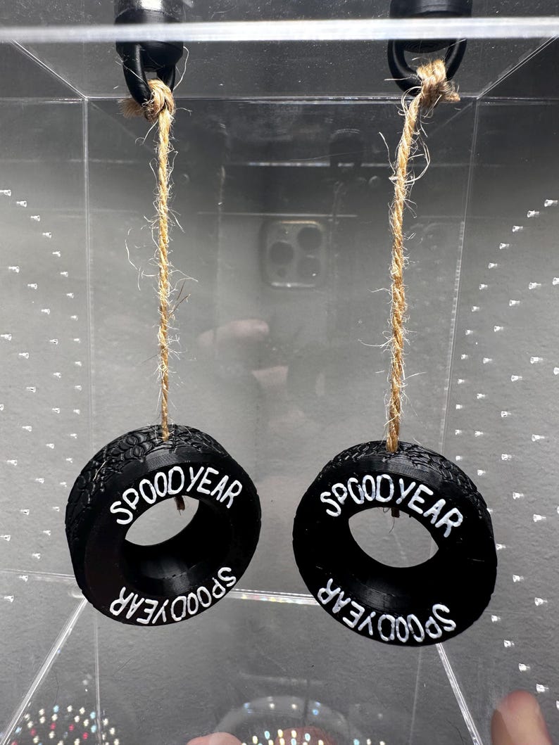 May include: Two black, tire-shaped ornaments with the text "SPOOD YEAR" in white lettering. Each ornament hangs from a natural-colored twine. The ornaments are suspended in front of a clear, reflective surface.